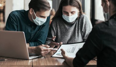 Maskeli iş arkadaşları ofis masasında oturuyorlar..