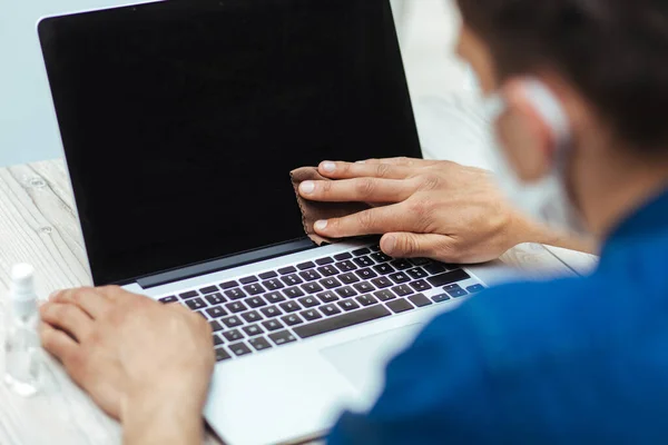 Close up. a young man in a protective mask wiping the screen of his laptop — Stock Photo, Image