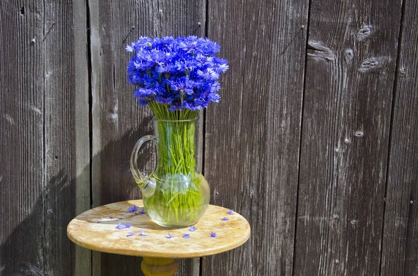 Cornflowers in glass jug on table near old wooden wall — Stock Photo, Image
