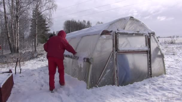 Jardineiro em limpeza vermelha removendo a neve da estufa de plástico — Vídeo de Stock