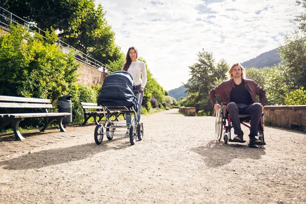 Young Parents In Wheelchair With Baby Stroller In The Park