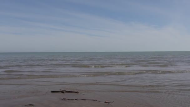 Песчаный пляж с парусником на горизонте, избитым спокойными волнами в летний день — стоковое видео