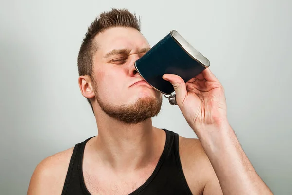 Young bearded guy drinks alcohol from a flask. On gray background. — Stock Photo, Image