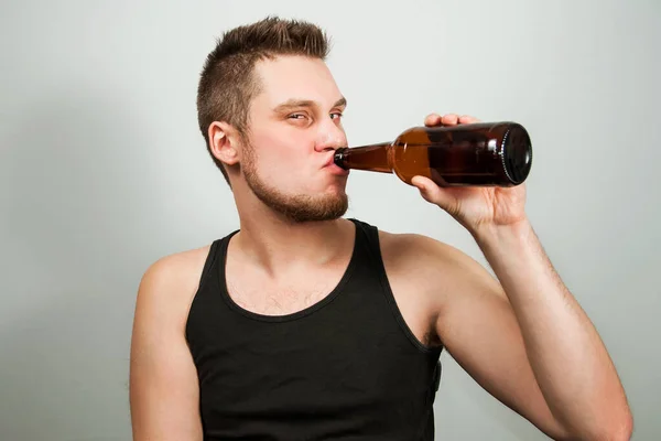 Junger Alkoholiker mit Kopfschmerzen, der eine Flasche Bier in der Hand hält. grauer Hintergrund. — Stockfoto