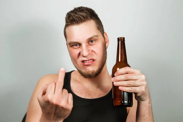 Dipendente giovane ragazzo alcolico mostra dito medio e tenendo in mano bottiglia di birra, su sfondo grigio . — Foto Stock