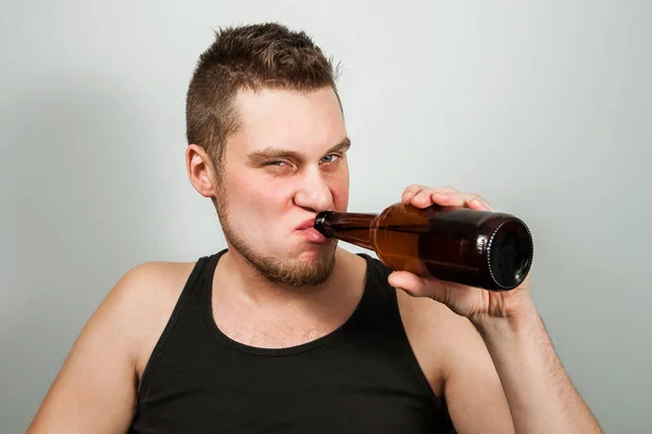 Junger Alkoholiker mit Kopfschmerzen, der eine Flasche Bier in der Hand hält. grauer Hintergrund. — Stockfoto