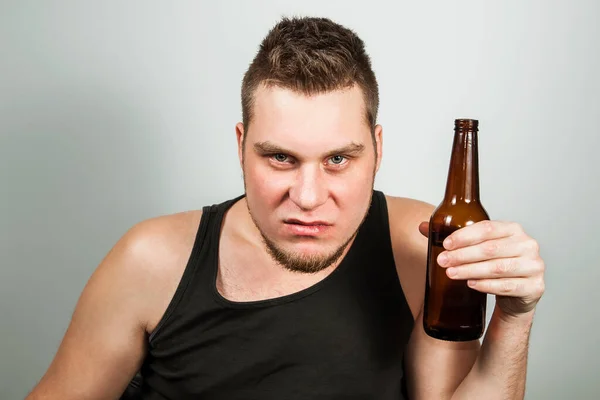 Young guy alcoholic with headache holding bottle of beer. Gray background. — Stock Photo, Image