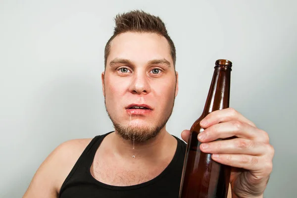 Betrunkener junger sabbernder Alkoholiker mit einer Flasche Bier auf grauem Hintergrund. — Stockfoto