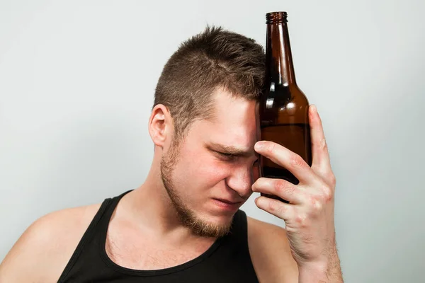 Junger Alkoholiker mit Kopfschmerzen, der eine Flasche Bier in der Hand hält. grauer Hintergrund. — Stockfoto