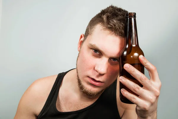 Junger Alkoholiker mit Kopfschmerzen, der eine Flasche Bier in der Hand hält. grauer Hintergrund. — Stockfoto