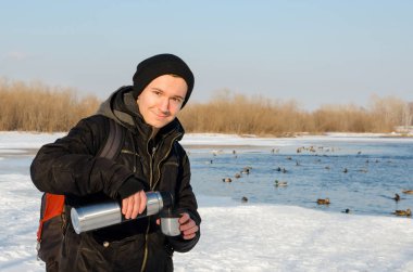 Ceketli genç adam, kış nehrinde ördekli termostan içiyor..
