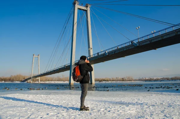 Mladý muž v bundě zahřívá ruce v zimě pod mostem řeka s kachnami pozadí. — Stock fotografie