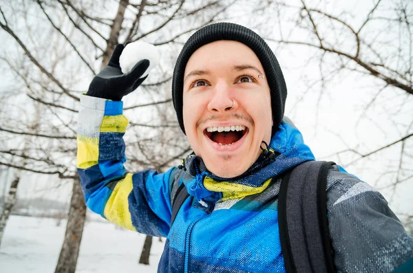Junger Mann mit Hut und Jacke wirft Schneeball auf Bäume. — Stockfoto