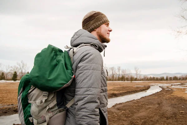 Junger Mann in grauer Jacke und Hut mit grünem Rucksack vor dem Hintergrund von Feld und Bäumen. — Stockfoto