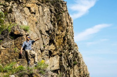 Genç bir turist tişört ve güneş gözlükleriyle kayaların arkasında seyahat eder.