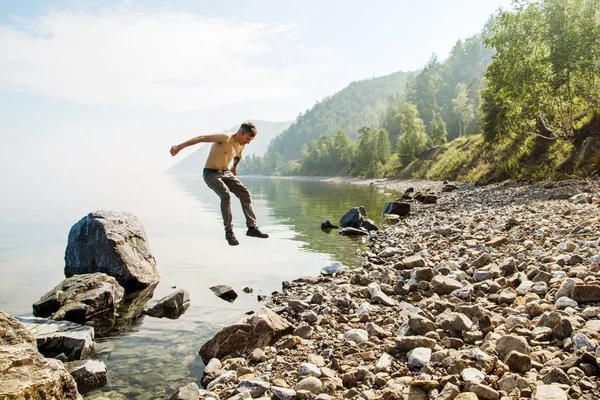 젊은 남자가 호수와 산을 배경으로 돌에서 뛰어 내립니다. — 스톡 사진