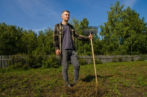 Ein junger Mann im Garten entfernt das abgeschrägte Gras mit einer Harke an einem Holzzaun und Waldhintergrund. — Stockfoto