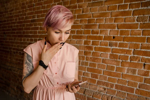 Joven hermosa chica sorprendida sostiene el teléfono inteligente sobre un fondo de pared de ladrillo — Foto de Stock