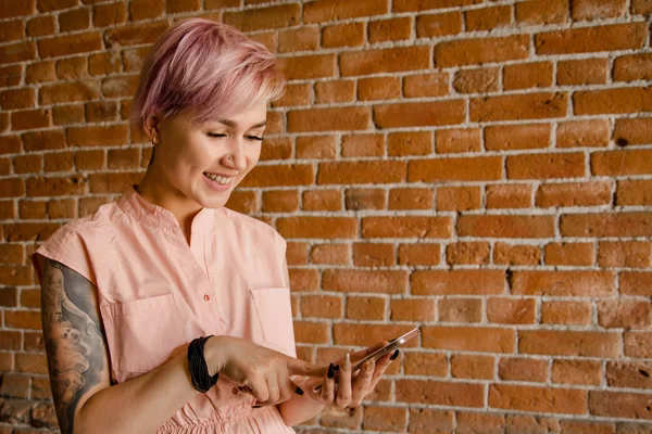 Joven hermosa chica sostiene el teléfono inteligente, leer y charlar sobre un fondo de pared de ladrillo — Foto de Stock