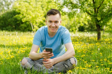 Genç adam cep telefonuyla iletişim kuruyor ve arka planda karahindiba ile yeşil çimlerin üzerinde oturuyor.