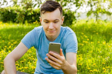 Genç adam akıllı telefondan iletişim kuruyor ve arka planda karahindiba ile yeşil çimlerin üzerinde oturuyor.