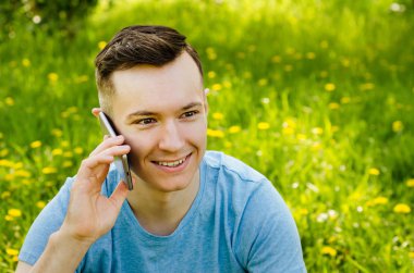 Genç adam akıllı telefondan iletişim kuruyor ve arka planda karahindiba ile yeşil çimlerin üzerinde oturuyor.