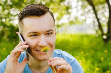 Genç adam akıllı telefondan iletişim kuruyor ve arka planda karahindiba ile yeşil çimlerin üzerinde oturuyor.