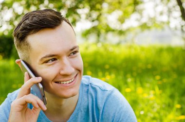Genç adam akıllı telefondan iletişim kuruyor ve arka planda karahindiba ile yeşil çimlerin üzerinde oturuyor.