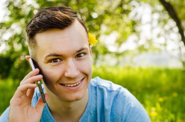 Genç adam akıllı telefondan iletişim kuruyor ve arka planda karahindiba ile yeşil çimlerin üzerinde oturuyor.