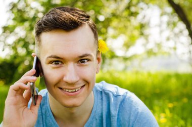 Genç adam akıllı telefondan iletişim kuruyor ve arka planda karahindiba ile yeşil çimlerin üzerinde oturuyor.