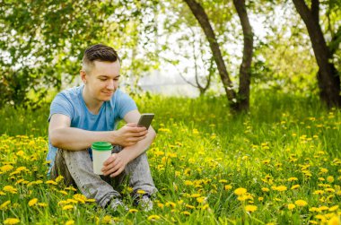 Genç adam cep telefonuyla iletişim kuruyor ve arka planda karahindiba ile yeşil çimlerin üzerinde oturuyor.