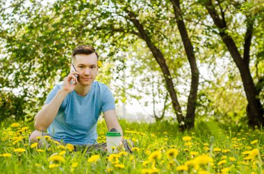 Genç adam akıllı telefondan iletişim kuruyor ve arka planda karahindiba ile yeşil çimlerin üzerinde oturuyor.