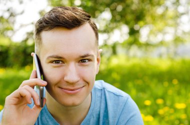 Genç adam akıllı telefondan iletişim kuruyor ve arka planda karahindiba ile yeşil çimlerin üzerinde oturuyor.