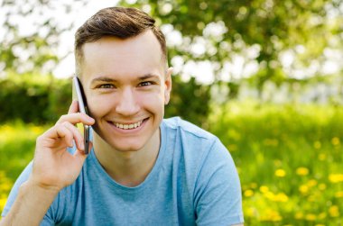 Genç adam akıllı telefondan iletişim kuruyor ve arka planda karahindiba ile yeşil çimlerin üzerinde oturuyor.