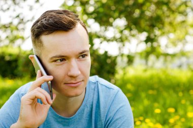 Genç adam akıllı telefondan iletişim kuruyor ve arka planda karahindiba ile yeşil çimlerin üzerinde oturuyor.