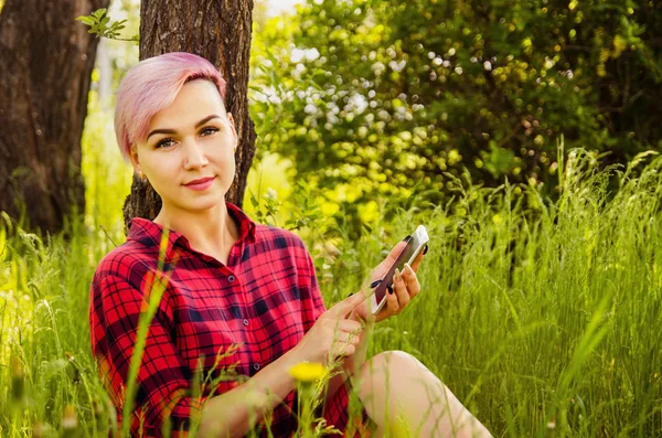 Joven hermosa chica sonriente con el pelo rosa sentado en la hierba verde, llamada por teléfono inteligente en la madera y el fondo del arbusto. Retrato. Mira en la cámara . — Foto de Stock