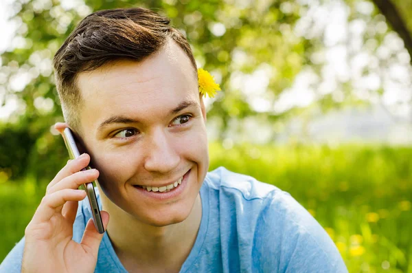 Genç adam akıllı telefondan iletişim kuruyor ve arka planda karahindiba ile yeşil çimlerin üzerinde oturuyor.