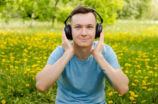 Portret młodego faceta słuchającego muzyki na słuchawkach i siedzącego na zielonej trawie z dmuchawcami. — Zdjęcie stockowe