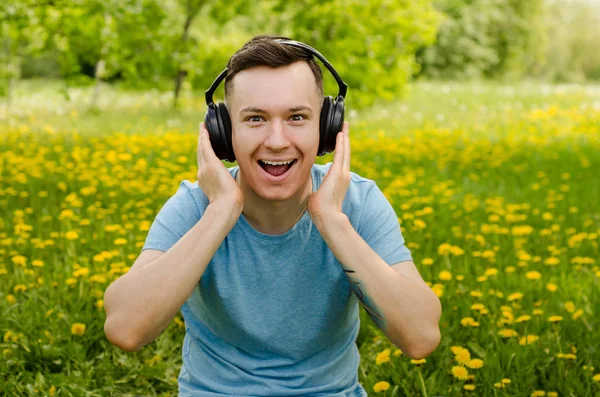 Portret van een jonge man die naar muziek luistert op een koptelefoon en op groen gras zit met paardebloemen. — Stockfoto
