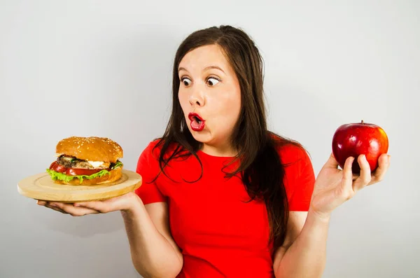Porträtt av en vacker fet flicka i överraskning och hålla ett rött äpple och en hamburgare, på grå bakgrund. — Stockfoto