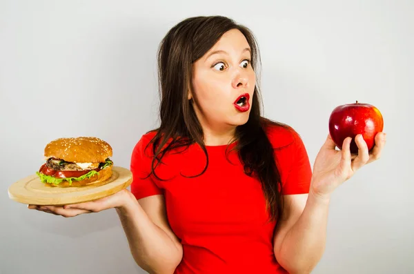 Porträtt av en vacker fet flicka i överraskning och hålla ett rött äpple och en hamburgare, på grå bakgrund. — Stockfoto