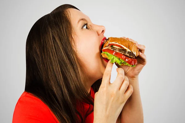 Wanita gemuk muda memegang hamburger, menatapnya dan tersenyum di latar belakang merah muda terang . — Stok Foto