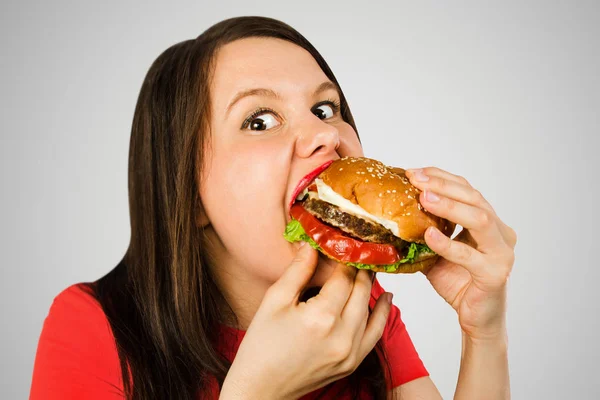 Young girl eats burger on gray background. Looks in camera. — 스톡 사진