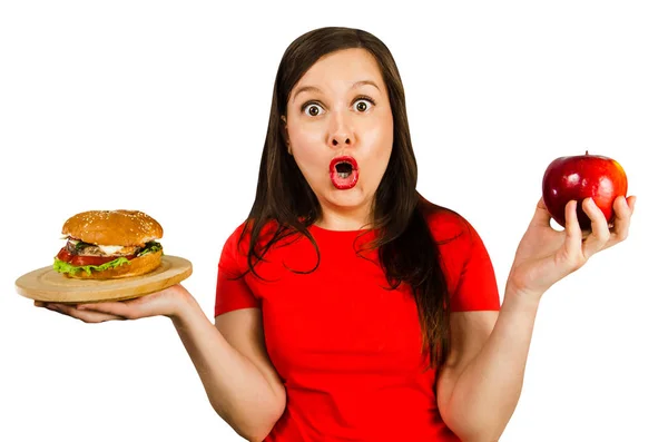 Portrait of beautiful fat girl holds burger and red apple and surprised with wide eyes. Isolated on white background. Stock Photo
