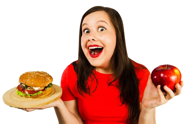 Güzel şişman kızın portresi hamburger ve kırmızı elma tutar ve geniş gözlerle şaşırır. Beyaz arkaplanda izole. Stok Fotoğraf