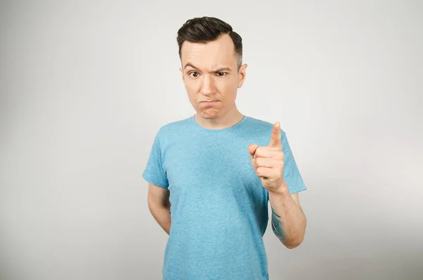 Νεαρός απαισιόδοξος άντρας με δάχτυλο, ντυμένος με μπλε μπλουζάκι σε ανοιχτό φόντο. Κλείσιμο πορτραίτου. — Φωτογραφία Αρχείου