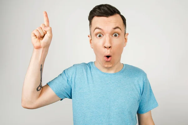 Ιδέα Νεαρός χαμογελαστός τύπος με το δάχτυλο ψηλά, ντυμένος με μπλε μπλουζάκι σε ανοιχτό φόντο. — Φωτογραφία Αρχείου