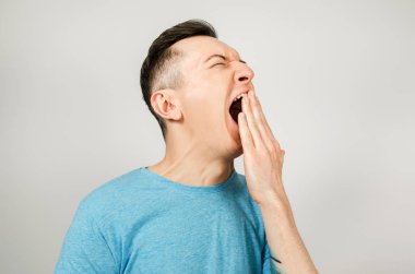 Açık arkaplanda mavi tişört giyen esneyen genç adam. Yan görünüm.