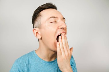 Açık arkaplanda mavi tişört giyen esneyen genç adam. Yan görünüm.