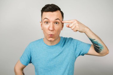 Mavi tişörtlü genç adam, arka planda parmağıyla şakağına işaret ediyor..